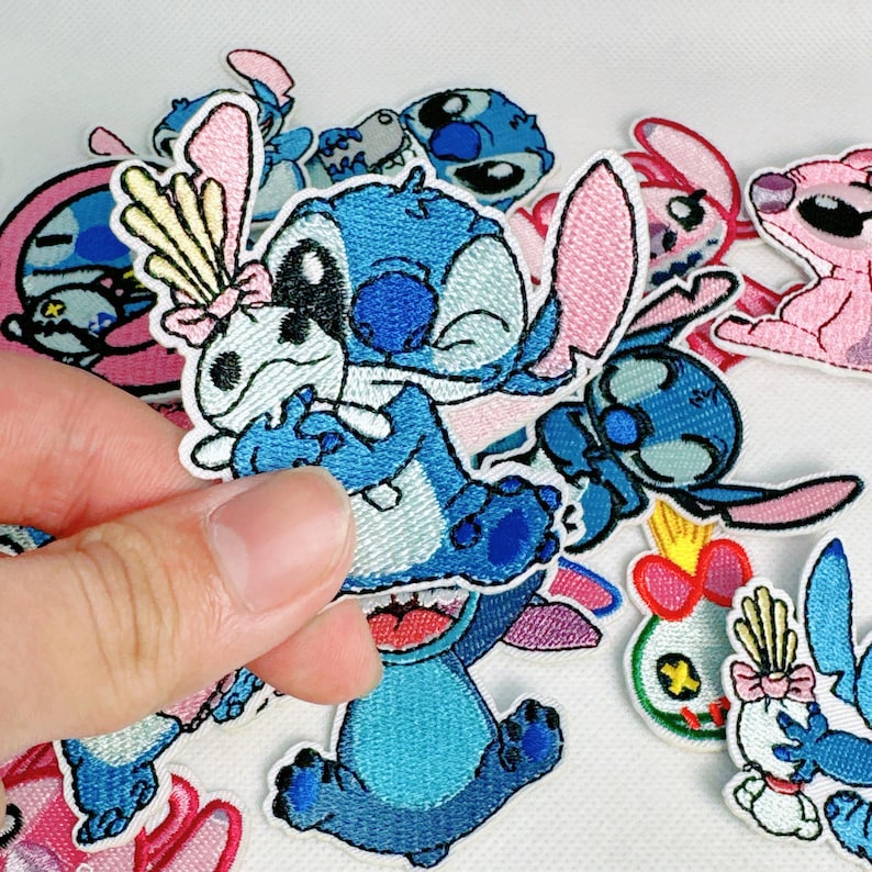 K&ouml;nnte beinhalten: Eine Sammlung von aufgestickten Aufn&auml;hern mit der Figur Stitch aus Lilo & Stitch. Die Aufn&auml;her sind haupts&auml;chlich blau, mit rosa und wei&szlig;en Akzenten. Einige Aufn&auml;her zeigen Stitch mit einer Schleife oder anderem Zubeh&ouml;r. Die Aufn&auml;her befinden sich auf wei&szlig;em Hintergrund.