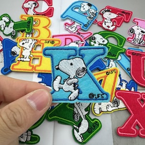 Può includere: Toppe ricamate con lettere dell'alfabeto e personaggi di Snoopy. Le toppe sono in vari colori, tra cui blu, verde, giallo e rosso. La lettera "K" è in primo piano, con Snoopy ricamato su uno sfondo azzurro.
