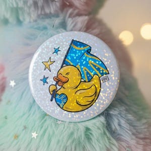 Könnte beinhalten: Ein runder, glitzernder Button mit einer gelben Ente, die eine blaue Flagge mit goldenen Sternen hält. Der Button ist an einem pastellfarbenen Plüschtier befestigt. Die Entenillustration hat einen lächelnden Ausdruck.