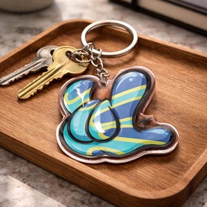 Könnte beinhalten: Ein klarer Acryl-Schlüsselanhänger mit einem abstrakten Design in Blau, Türkis und Gelb. Der Schlüsselanhänger ist an einem silbernen Schlüsselring mit einer Kette befestigt und liegt auf einem Holztablett mit Schlüsseln.