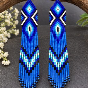 May include: A pair of long, dangling earrings crafted from small glass beads in shades of royal blue, light blue, white, and black. The design features a geometric diamond pattern at the top and a chevron-style fringe at the bottom with silver hooks.