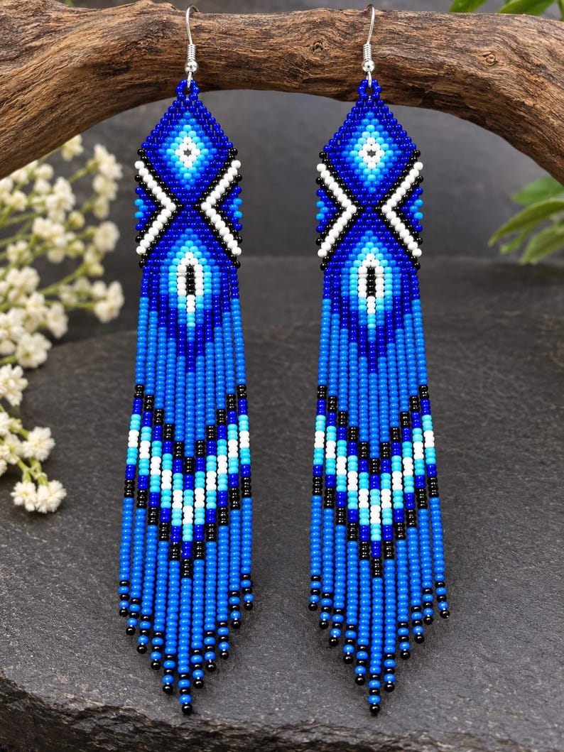 May include: A pair of long, dangling earrings crafted from small glass beads in shades of royal blue, light blue, white, and black. The design features a geometric diamond pattern at the top and a chevron-style fringe at the bottom with silver hooks.