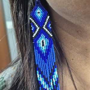 May include: Long, dangling beaded earrings featuring a geometric pattern in shades of royal blue, light blue, white, and black. The design consists of small, uniform glass beads arranged in a diamond motif with a fringe of vertical beaded strands.