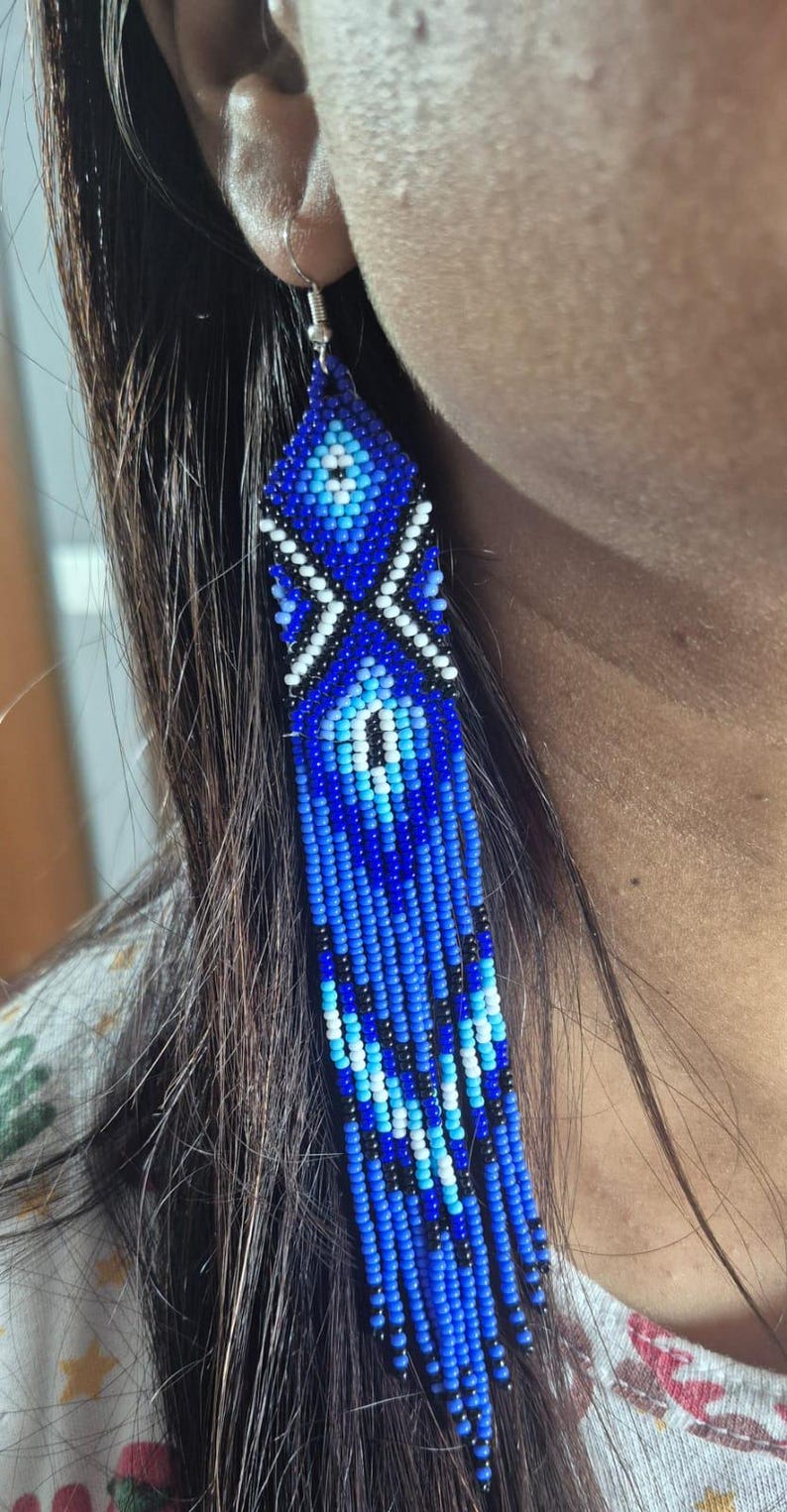 May include: Long, dangling beaded earrings featuring a geometric pattern in shades of royal blue, light blue, white, and black. The design consists of small, uniform glass beads arranged in a diamond motif with a fringe of vertical beaded strands.