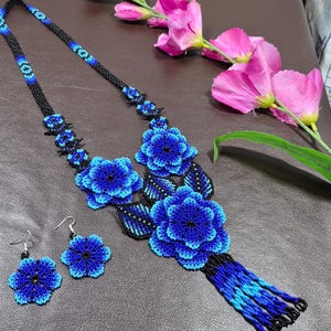 May include: A beaded necklace and matching earring set featuring intricate floral designs in shades of blue and black. The necklace includes a long beaded chain, two large three-dimensional blue flower pendants, black beaded leaves, and a blue fringe.