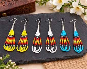 May include: Three pairs of dangling earrings featuring multi-colored seed beads arranged in vertical strands. The beads are colored in yellow, white, and turquoise, each accented with horizontal bands of red, orange, yellow, and black, with silver-tone cones.