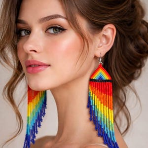 May include: Long, triangular dangle earrings featuring a vibrant rainbow pattern made of small, multi-colored seed beads. The design includes a horizontal black band near the top, with long, cascading beaded fringe strands in red, orange, yellow, green, and blue.