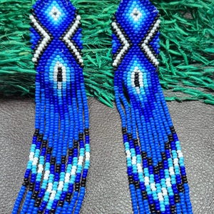 May include: A pair of long, dangling earrings crafted from small seed beads in shades of royal blue, light blue, white, and black. The earrings feature a geometric diamond pattern at the top and a long, multi-strand fringe design at the bottom.