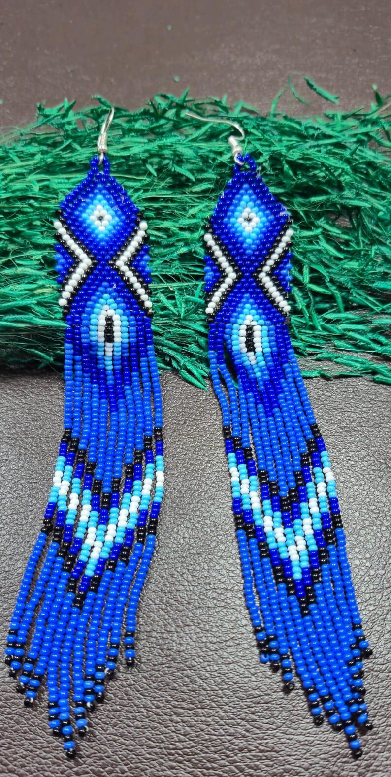 May include: A pair of long, dangling earrings crafted from small seed beads in shades of royal blue, light blue, white, and black. The earrings feature a geometric diamond pattern at the top and a long, multi-strand fringe design at the bottom.