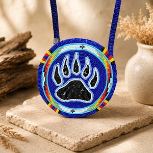 May include: A circular medallion necklace featuring a black bear paw print design crafted from small blue, black, and white beads. The outer edge is decorated with a vibrant border of yellow, red, and white beads, attached to a blue beaded strap.