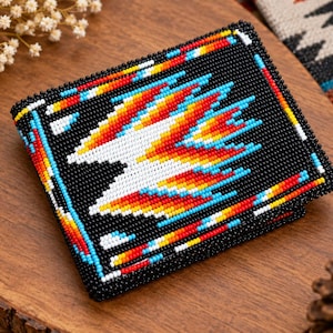 May include: A square wallet featuring intricate beadwork with a black background and a central geometric pattern in white, orange, yellow, and light blue. The edges are bordered by a colorful beaded frame of red, yellow, and blue accents.