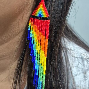 May include: A long, dangling earring crafted from small, vibrant seed beads arranged in a rainbow pattern. The design features a triangular top section with a horizontal black band, followed by multiple cascading fringe strands in red, orange, yellow, green, light blue, and dark blue.