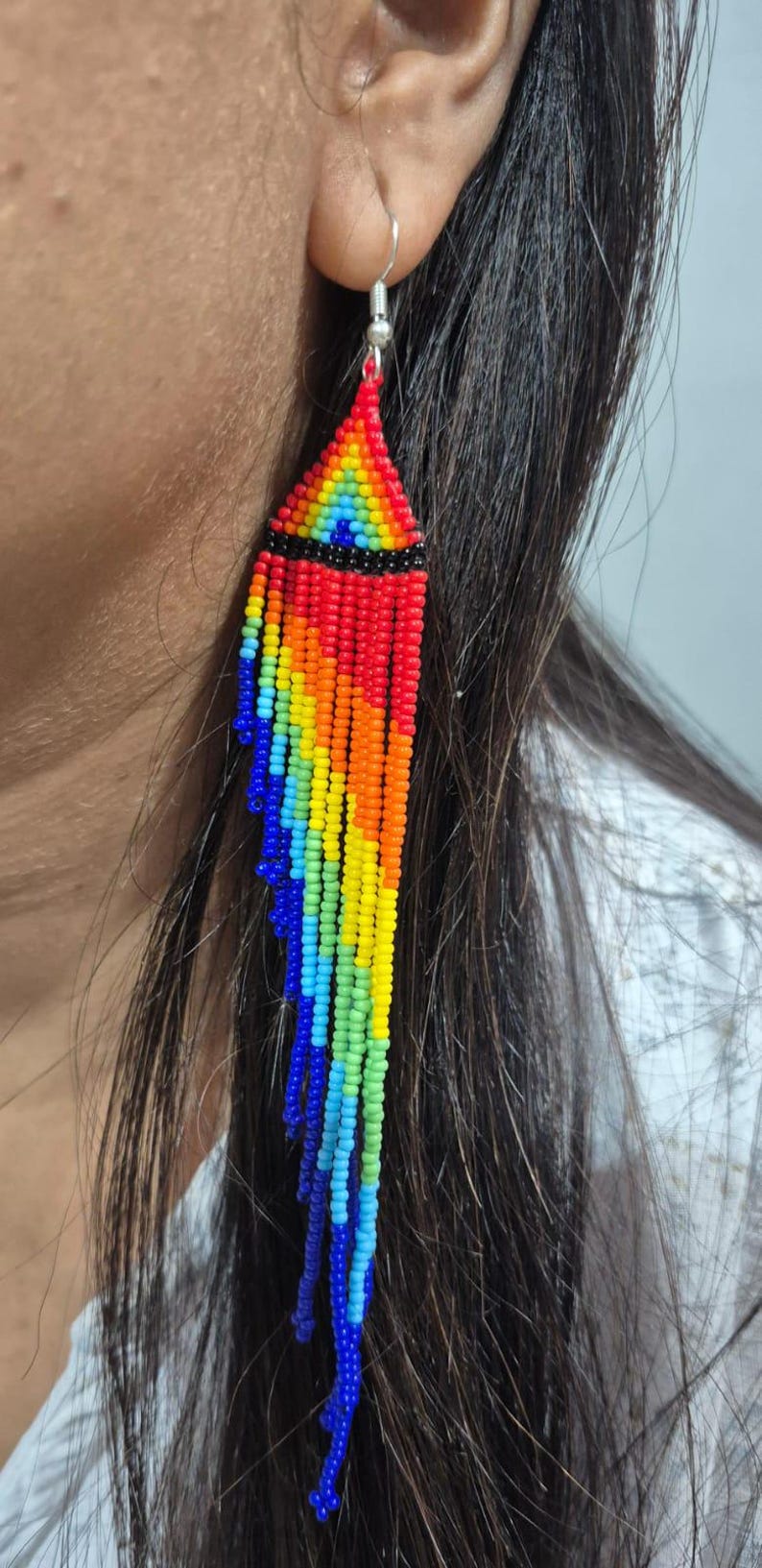 May include: A long, dangling earring crafted from small, vibrant seed beads arranged in a rainbow pattern. The design features a triangular top section with a horizontal black band, followed by multiple cascading fringe strands in red, orange, yellow, green, light blue, and dark blue.