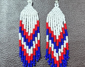 May include: A pair of long, dangling earrings crafted from small, spherical beads in white, red, and blue. The beads are arranged in a chevron pattern that transitions into a fringe at the bottom. Each earring features a silver-toned metal hook.