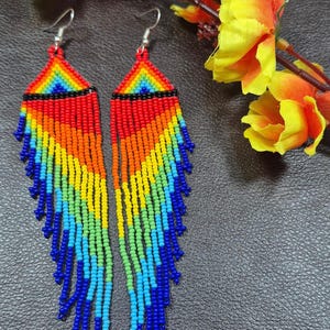 May include: A pair of long, dangling earrings crafted from small, multi-colored seed beads arranged in a geometric rainbow pattern. The design features a triangular top section with horizontal stripes of red, orange, yellow, green, and blue, followed by long, cascading fringe strands in a matching spectrum of vibrant colors.