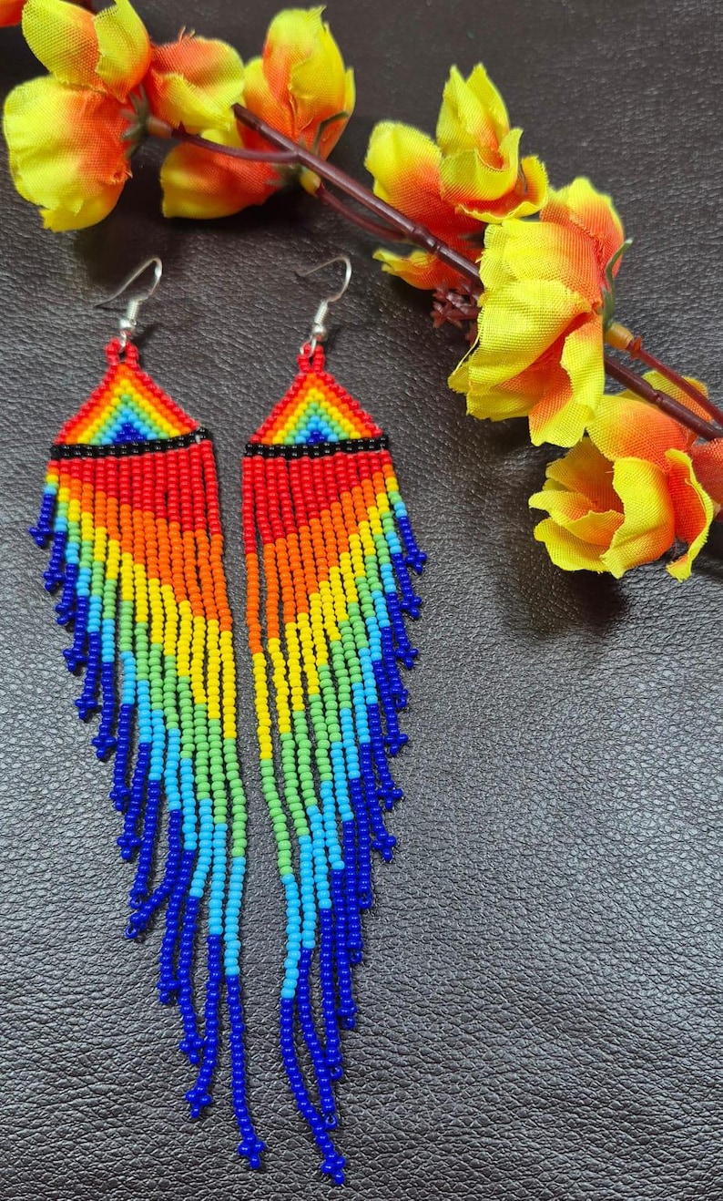 May include: A pair of long, dangling earrings crafted from small, multi-colored seed beads arranged in a geometric rainbow pattern. The design features a triangular top section with horizontal stripes of red, orange, yellow, green, and blue, followed by long, cascading fringe strands in a matching spectrum of vibrant colors.