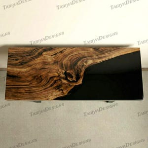 May include: A rectangular coffee table with a live edge wood top and a black resin inlay. The wood features a natural grain pattern in shades of brown and tan. The table has metal legs.