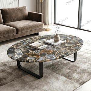 Puede incluir: Mesa de centro ovalada con una tapa de piedra de mosaico en tonos grises, marrones y blancos. La mesa tiene una base de metal negro y está sobre una alfombra de tonos neutros. Un pequeño jarrón y una bandeja con libros están sobre la mesa.