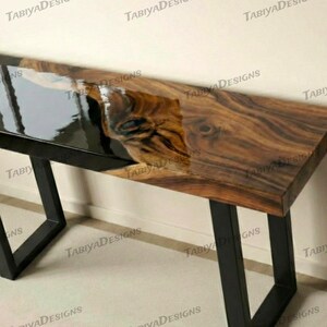 May include: A wooden console table with a live edge design. It has a dark, glossy epoxy resin inlay and black metal legs. The wood grain is visible, displaying natural patterns. The table's length is approximately 150 cm.