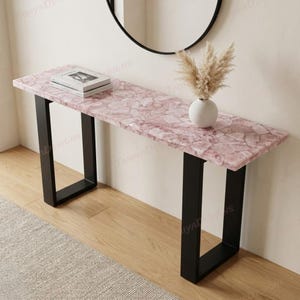May include: A rectangular console table featuring a pink quartz stone top with a mosaic-like texture and two black metal U-shaped legs. A white vase containing dried pampas grass and a stack of two books are placed on the surface of the table.