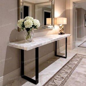 May include: A white marble console table with black metal legs. A clear glass vase with white flowers sits on the table. A gold lamp with a cream shade is on the table. A mirror hangs above the table.