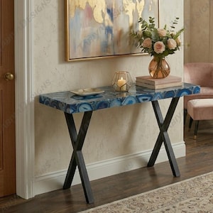May include: A rectangular console table featuring a blue agate stone top with natural banding patterns and a black metal X-shaped base. The table surface holds a small glass candle holder with a white candle and a stack of two books.