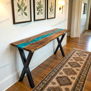 Puede incluir: Una consola de madera con un diseño de río de resina turquesa. La mesa tiene un acabado de vetas de madera natural y patas de metal negro en forma de X. La mesa mide aproximadamente 183 cm de largo.