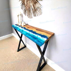 May include: A handcrafted console table with a wooden top and a blue resin design resembling ocean waves. The table has black metal legs and is decorated with candles and decorative objects. The table measures approximately 48 inches long.