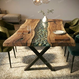 May include: A handcrafted wooden dining table with a river of clear resin filled with pebbles. The table has a natural wood grain finish and a black metal base. The table is approximately 72 inches long.
