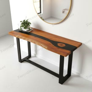 May include: A wooden console table with a live edge design and a black epoxy resin inlay. The table has a natural wood grain finish and sits on black metal legs. A small plant and books are on top.