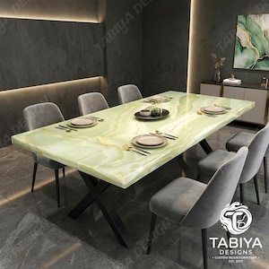 Puede incluir: Una mesa de comedor rectangular con una superficie pulida de patrón de ónix verde claro y una base de metal negro en forma de X. La mesa está dispuesta con cuatro sillas tapizadas en gris, cada una con respaldo redondeado y patas finas.
