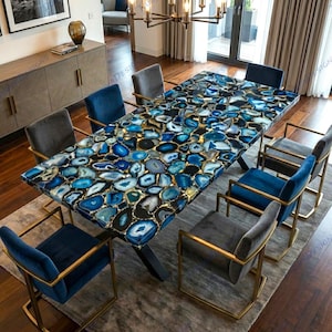 May include: A rectangular dining table with a blue agate stone top and a black base. The table is surrounded by eight chairs with gold-colored frames and blue or gray velvet seats. The table is approximately 100 inches long.