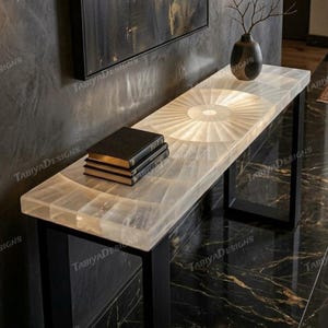 May include: A rectangular console table with a geometric design on the top surface. The table has a light-colored, possibly translucent, top with a sunburst pattern. It rests on black metal legs. Three books are stacked on the table.