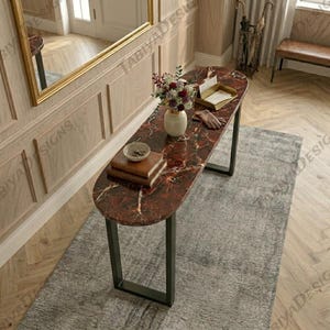 May include: A long, oval-shaped console table with a dark red, marble-patterned top and black metal U-shaped legs. On the surface sits a white ceramic vase filled with flowers, a stack of two brown books, and a small gold-toned tray with paper.