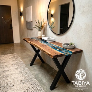 May include: A console table featuring a natural live-edge wood slab with a central river inlay of blue resin and embedded river stones. The table is supported by black metal X-shaped legs and displays the Tabiya Designs logo in the corner.