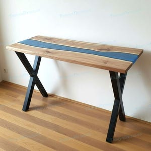 May include: A wooden desk with a blue resin river design. The desk has a rectangular top with a light brown wood grain and a blue center. Black metal legs provide support. The desk is approximately 48 inches long.