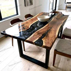 May include: A rectangular dining table with a live edge design. The table features a dark epoxy river running through the center, framed by natural wood. The base is black metal. A white bowl sits on the table.
