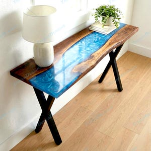 May include: A wooden console table with a blue resin river design. The table has a dark wood border and black metal legs. A white lamp and a small potted plant sit on the table.