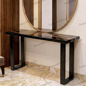 May include: A modern console table with a dark, glossy top and black legs. The table is positioned against a wall with a large, round mirror and vertical wood paneling. The text "TABIYA DESIGNS" is visible.