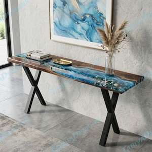 May include: A long console table featuring a natural wood slab with a central blue resin river inlay containing embedded stones. The table is supported by black metal X-shaped legs and holds a glass vase with dried grasses and a stack of books.