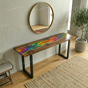 May include: A wooden console table with a vibrant, multicolored resin river design. The table has a dark wood finish and black metal legs. A round gold mirror hangs on the wall above the table. A small potted tree is to the right.