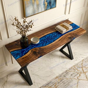May include: A handcrafted wooden table with a vibrant blue resin river running through the center. The table features a dark wood grain, black metal legs, and is adorned with decorative items, including a vase and a bowl.