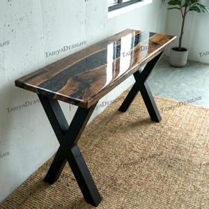 May include: A rectangular wooden desk with a dark epoxy resin inlay. The tabletop is supported by black, X-shaped metal legs. The desk is set on a woven jute rug. The brand name "TabiyaDesigns" is printed on the desk.