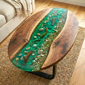 Puede incluir: Mesa de centro ovalada con un diseño único. La mesa presenta un río de resina transparente de color turquesa lleno de piedras y guijarros. Los bordes son de madera natural y tiene una base de metal negro.
