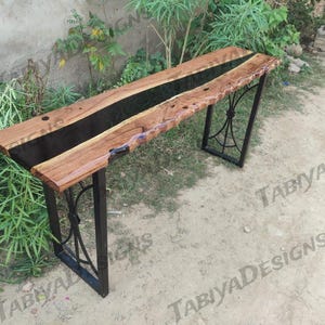 Puede incluir: Un escritorio de madera hecho a mano con un centro de resina epoxi oscura. El escritorio presenta un acabado de vetas de madera natural y está sostenido por patas de metal negro con detalles decorativos. El diseño general es rústico y moderno.