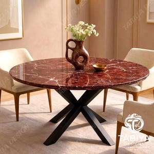 Op de afbeelding: Een ronde eettafel met een gepolijst dieprood marmeren blad met witte aders, ondersteund door een zwart metalen X-vormig onderstel. De tafel is omringd door drie crèmekleurige gestoffeerde stoelen en bevat een bruine keramische vaas met witte bloemen.