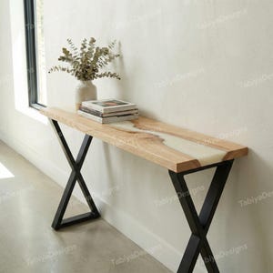May include: A wooden console table with a light wood top and a white resin inlay. The table has black metal X-shaped legs. A vase with dried branches and a stack of books are on the table.