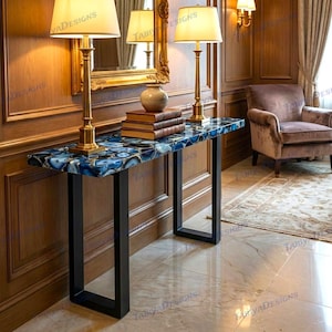 May include: A rectangular console table with a blue and gold agate top and black metal legs. Two gold lamps with cream-colored shades sit on the table, along with a stack of books and a decorative vase. The table is placed against a wood-paneled wall.