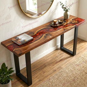 May include: A long, wooden console table with a striking red resin river design. The table has a dark wood finish and black metal legs. A book, a tray with a candle, and a vase with greenery are on top of the table.