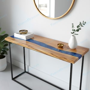 May include: A wooden console table with a blue resin river design. The table has a rectangular top and black metal legs. A gold-colored bowl, a stack of books, and a white vase with greenery are on the table. A gold-framed mirror is in the background.